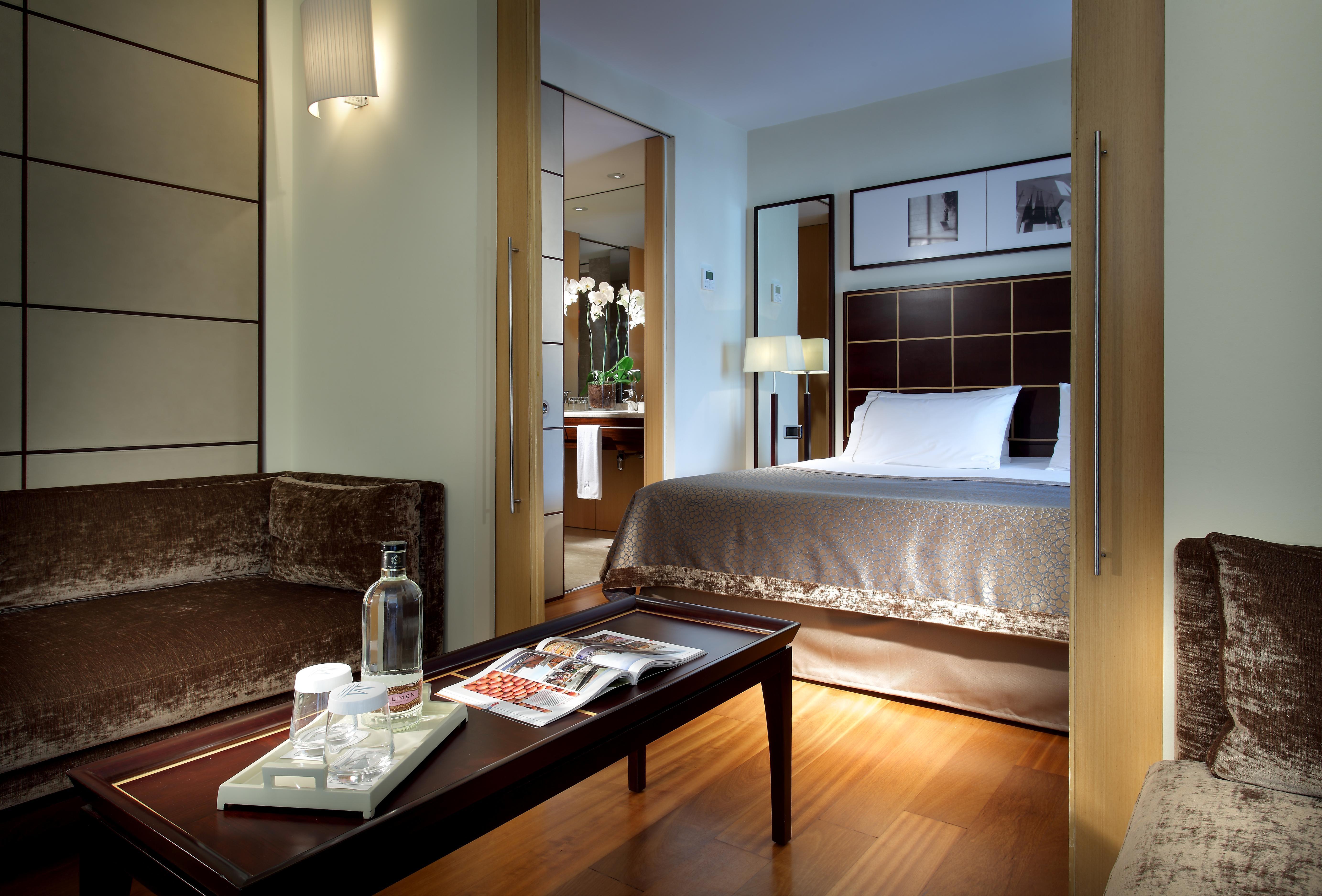 Eurostars Grand Marina Hotel Gl Barcelona Bagian luar foto The image shows a well-decorated hotel room featuring a comfortable bed with a plush headboard, complemented by a subtle color palette. There is a cozy seating area with a brown couch and a coffee table. On the table, there are items like a water bot