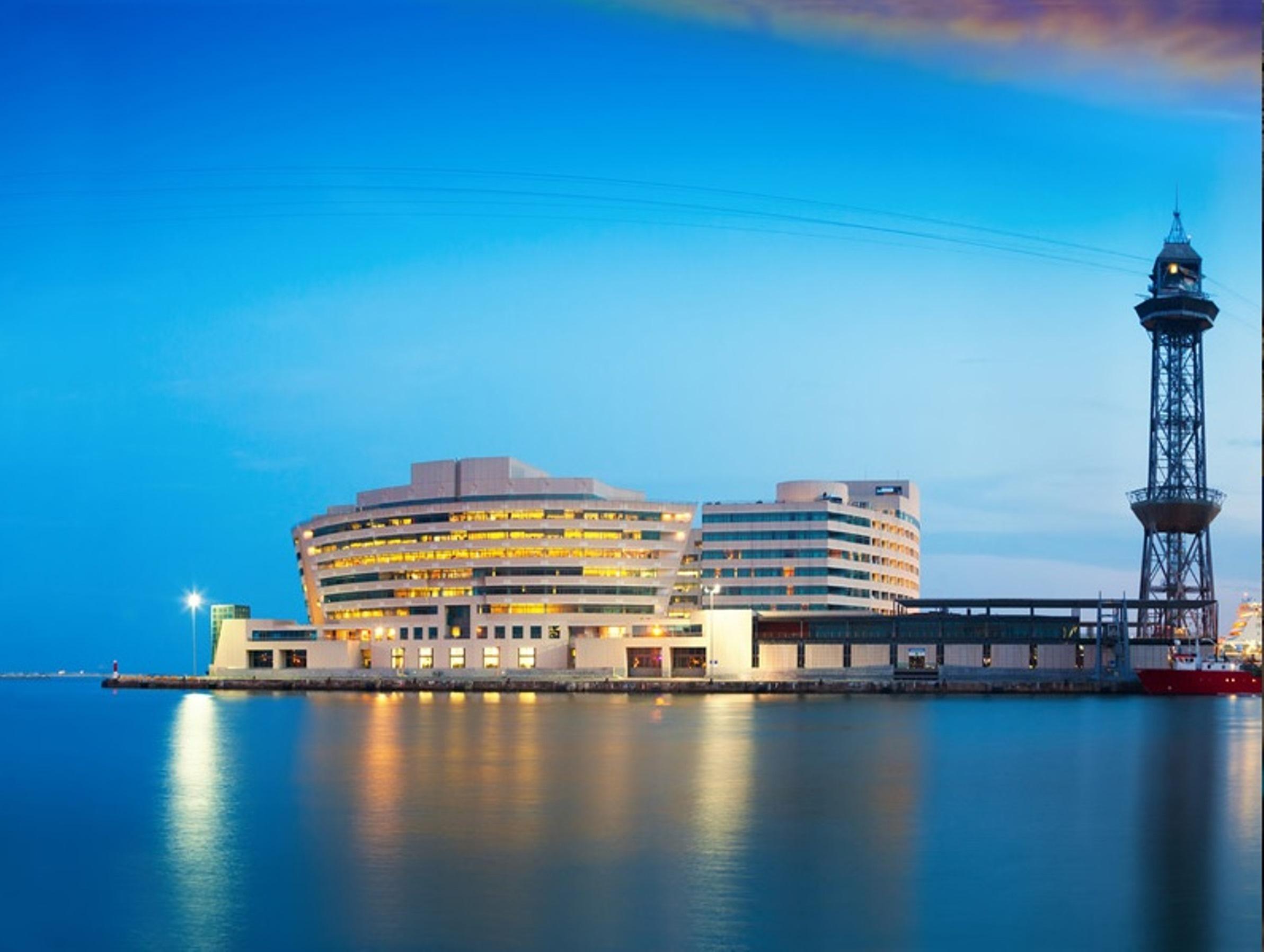 Eurostars Grand Marina Hotel Gl Barcelona Bagian luar foto The photo shows a modern building situated near the water, likely a harbor or port area. The structure features a sleek, curved design with multiple levels and large windows, reflecting an urban architectural style. Next to the building, there is a t