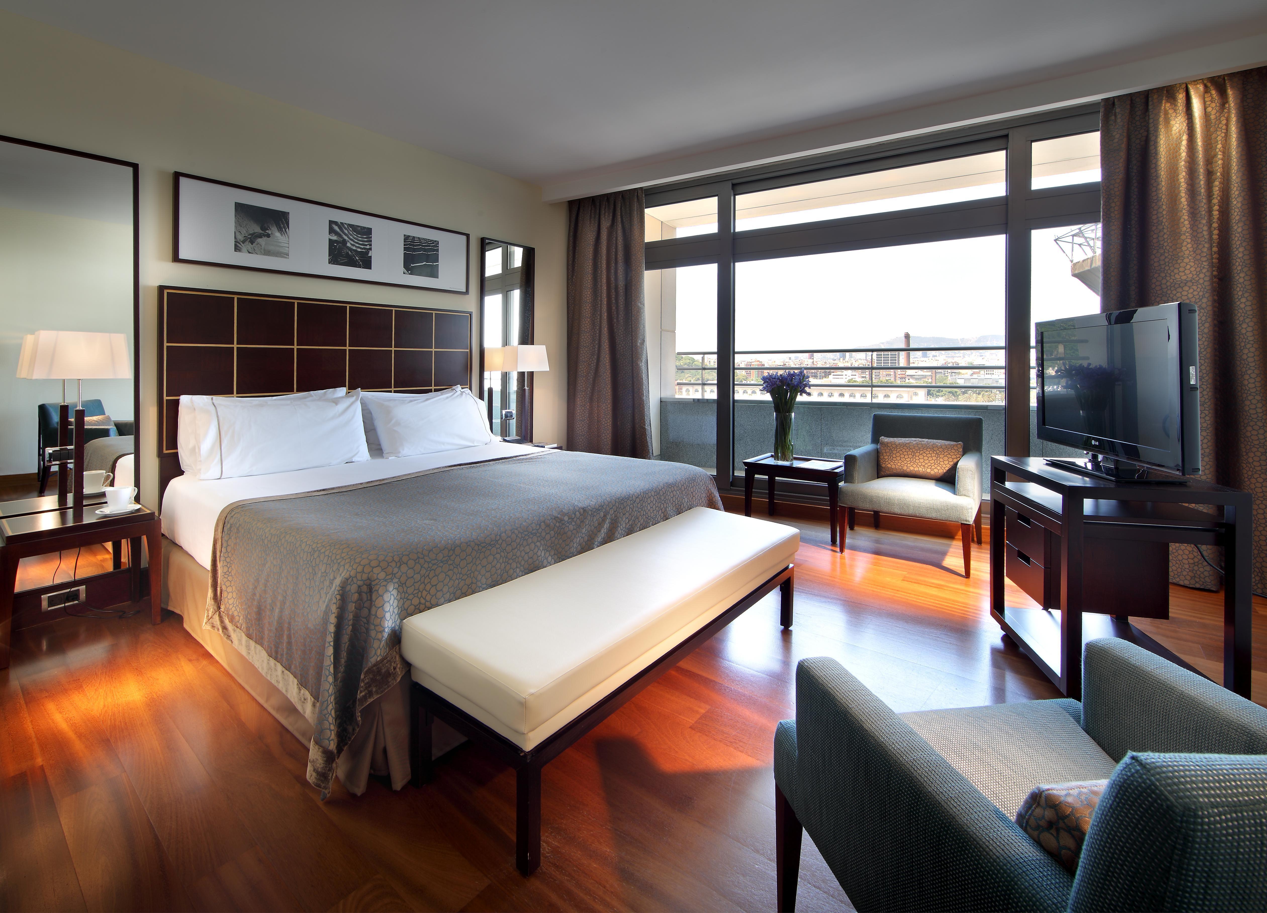 Eurostars Grand Marina Hotel Gl Barcelona Bagian luar foto The photo shows a cozy hotel room featuring a large bed with white linens and a gray bedspread. There is a light-colored bench at the foot of the bed. A small seating area includes a comfortable chair and a lamp, along with a wooden side table. The r
