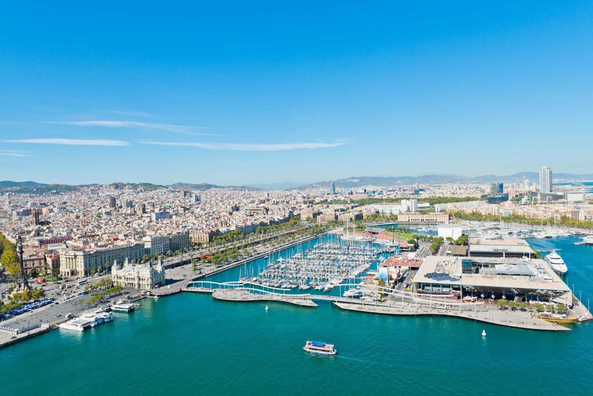 Eurostars Grand Marina Hotel Gl Barcelona Bagian luar foto The photo depicts a coastal city view, likely of a vibrant harbor or marina. In the foreground, there is a clear blue body of water with a few small boats. The harbor features numerous yachts and boats docked in the marina. 

Surrounding the water ar