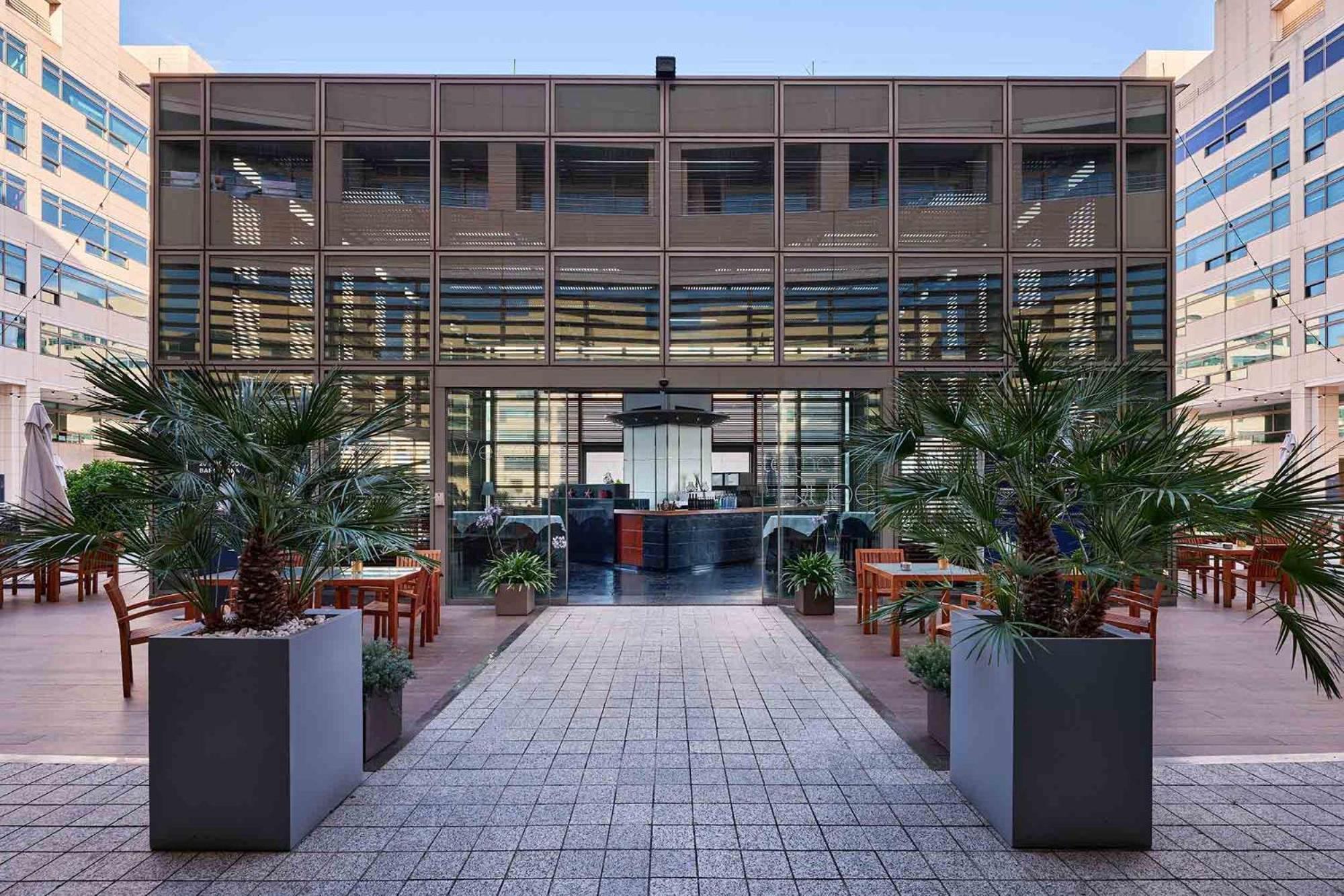 Eurostars Grand Marina Hotel Gl Barcelona Bagian luar foto The photo depicts a modern building with a glass facade, featuring a central entrance framed by large planters with tropical plants. The entrance leads to an interior space that appears to be a restaurant or café, with a stylish counter and seating v