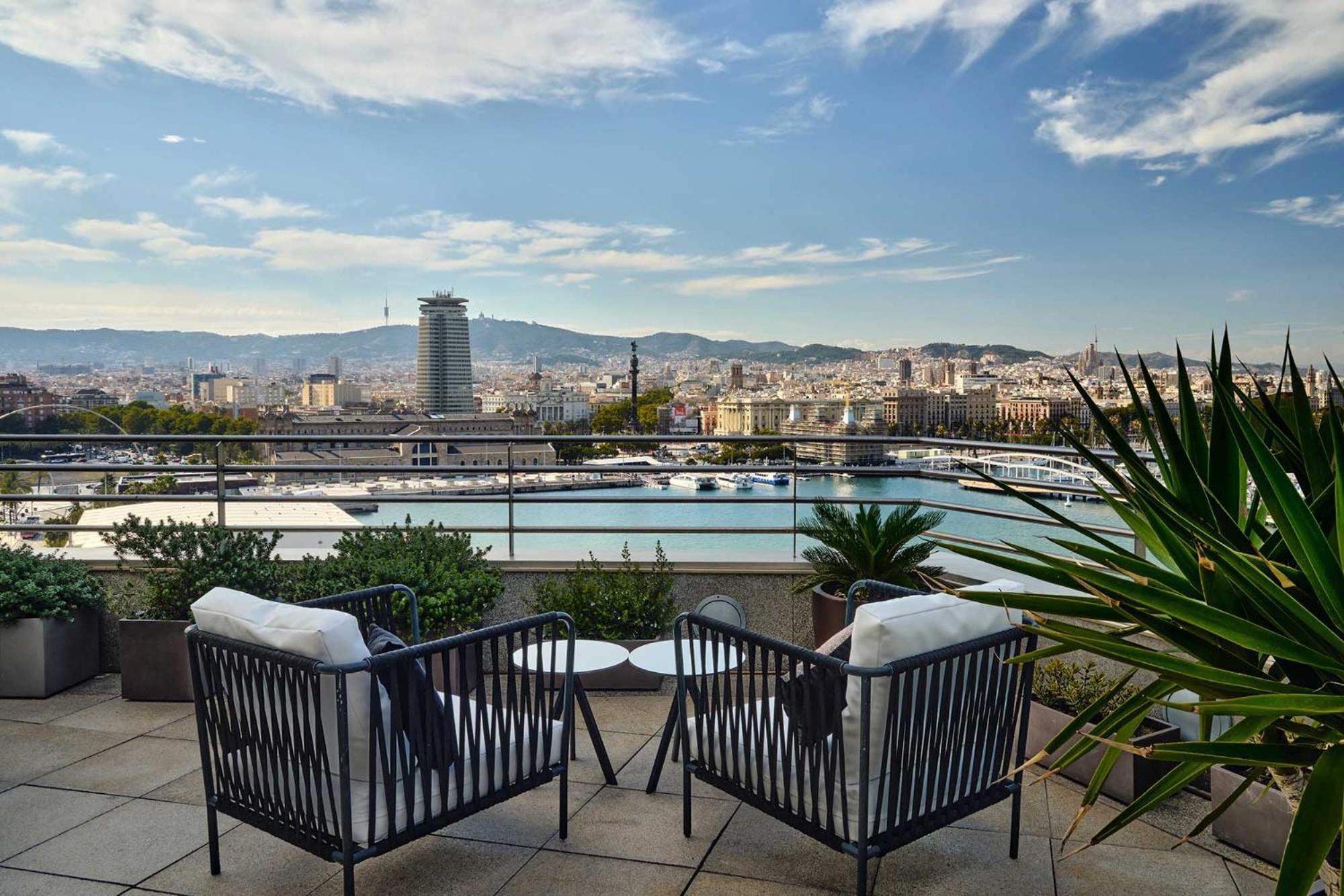 Eurostars Grand Marina Hotel Gl Barcelona Bagian luar foto The photo shows a scenic outdoor terrace area featuring two stylish black chairs with white cushions. In the background, there’s a beautiful view of a cityscape with a harbor and mountains in the distance. The sky is partly cloudy, and there are some