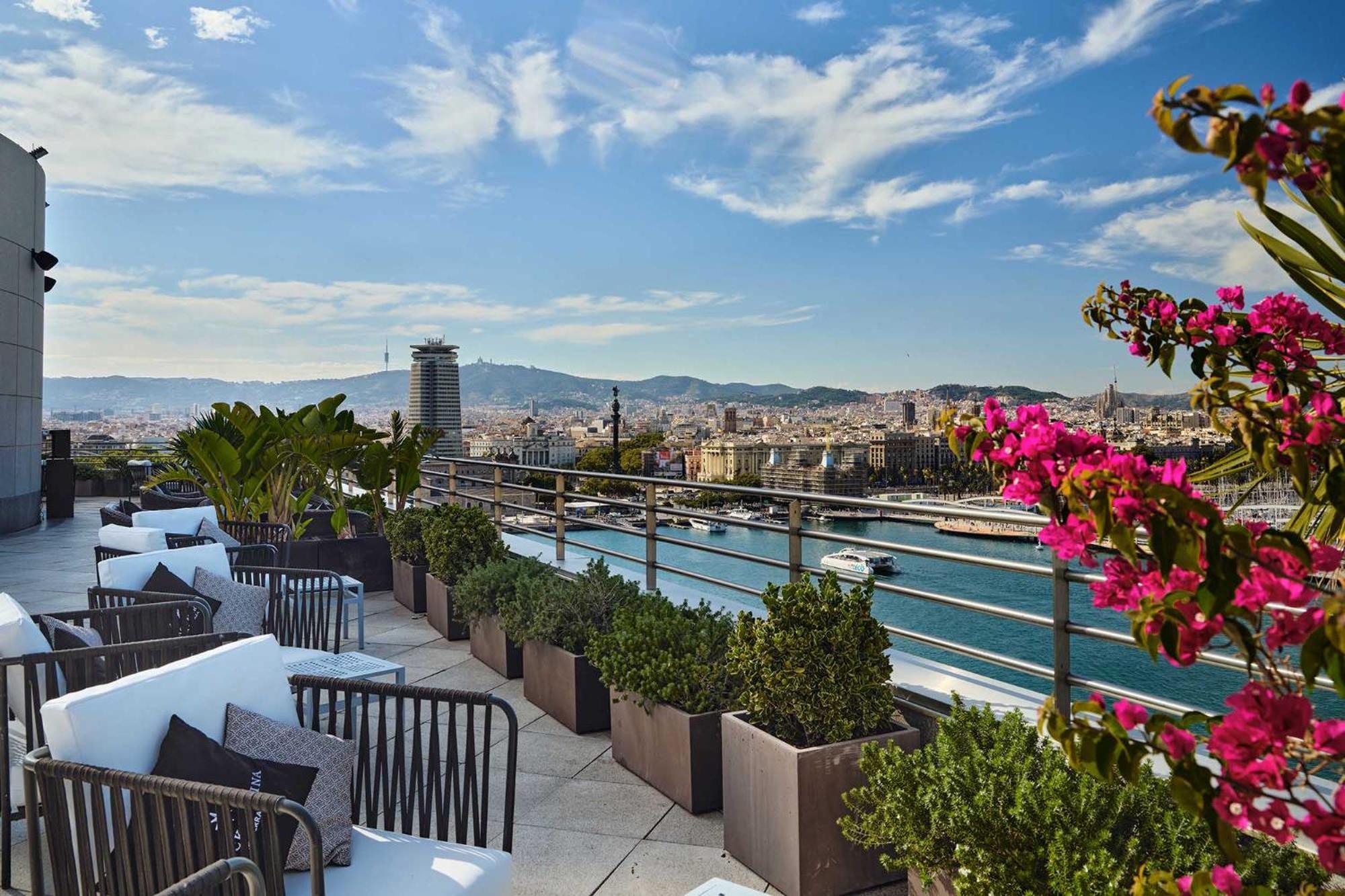 Eurostars Grand Marina Hotel Gl Barcelona Bagian luar foto The photo depicts a rooftop terrace with a stunning view of a city, likely Barcelona. There are comfortable seating arrangements, including dark outdoor chairs and cushions, surrounded by decorative plants in large pots. Colorful flowers, possibly bo