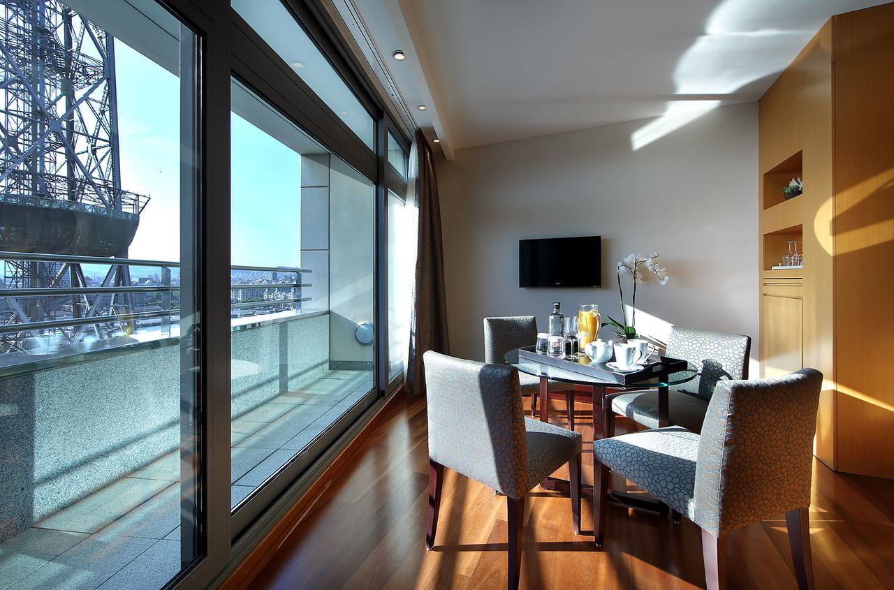 Eurostars Grand Marina Hotel Gl Barcelona Bagian luar foto The photo shows a modern and bright room that features large windows with panoramic views. Inside, there is a circular dining table surrounded by upholstered chairs. On the table, there are items that suggest a meal or refreshments, such as drinks an