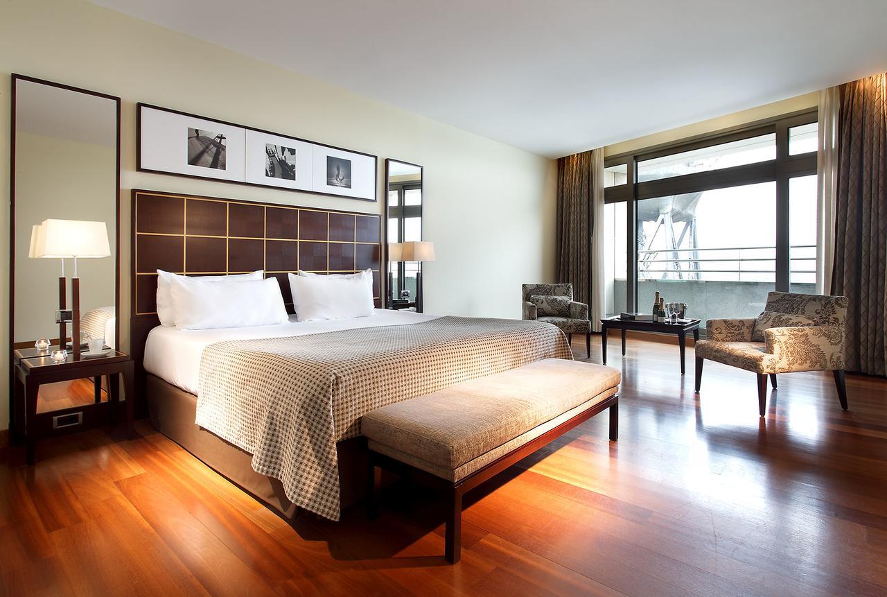Eurostars Grand Marina Hotel Gl Barcelona Bagian luar foto The photo shows a modern hotel room featuring a large bed with a decorative headboard. There are two bedside lamps on either side of the bed, and behind the bed, there are framed pictures hanging on the wall. A bench sits at the foot of the bed, and 