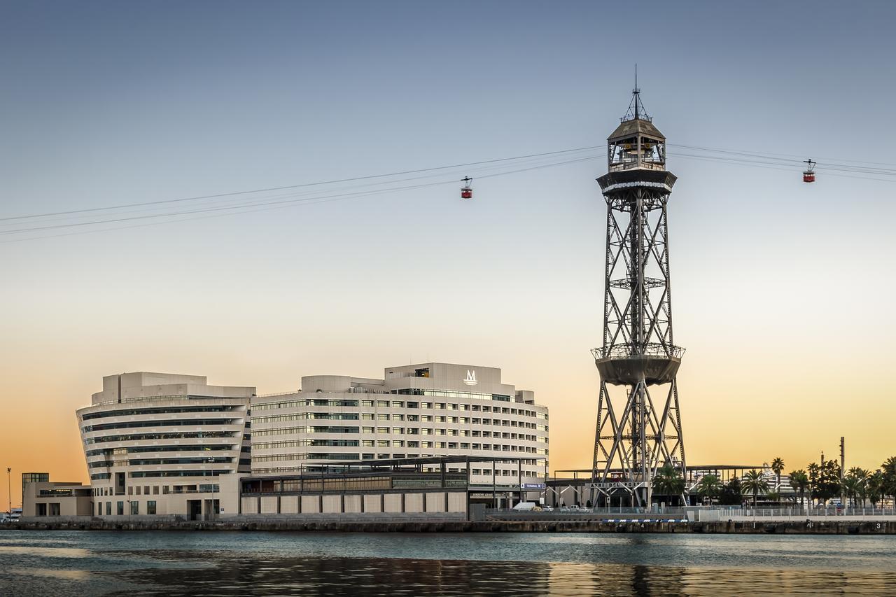 Eurostars Grand Marina Hotel Gl Barcelona Bagian luar foto The photo shows a modern building alongside a waterfront, with a distinctive tower nearby. The tower appears to be part of a cable car system, featuring a conical roof and a series of cables extending from it. The scene is set against a colorful sky,